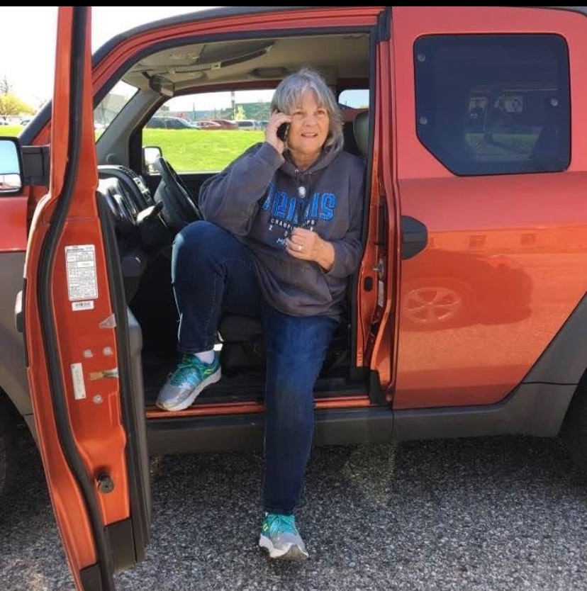 Rosemary exiting the driver side of a vehicle while talking on a cell phone and looking past the camera.